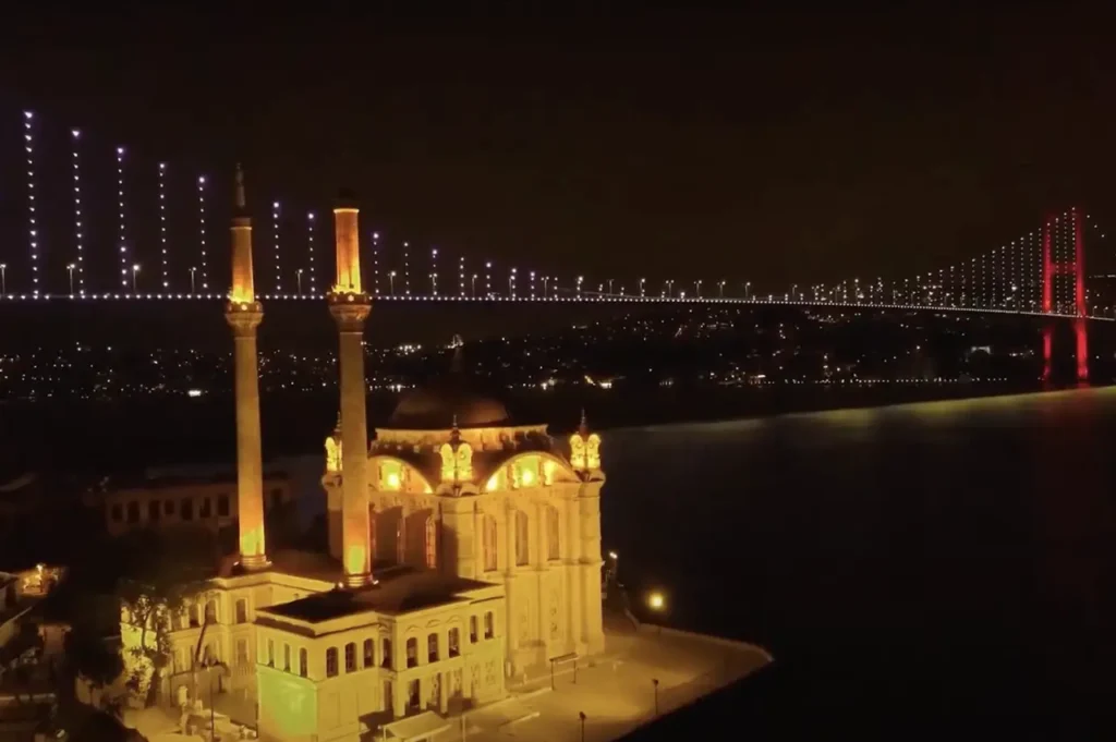 The Bosphorus Bridge illuminated at night, viewed from Ortaköy, with bright lights reflecting on the water.