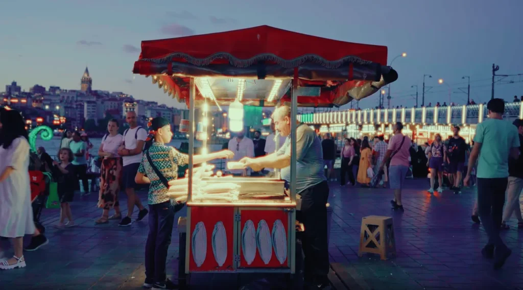 Corn vendor near Galata Bridge with a red and white cart, and views of the bustling waterfront.